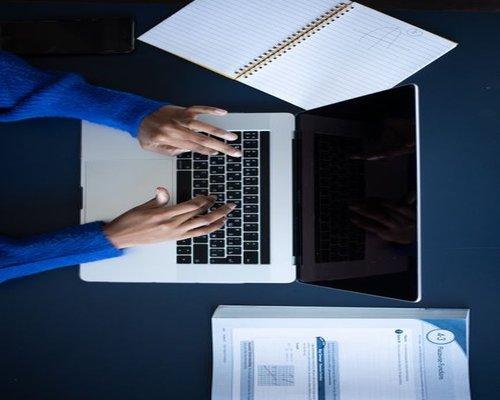 Hands typing on a laptop with a notebook nearby
